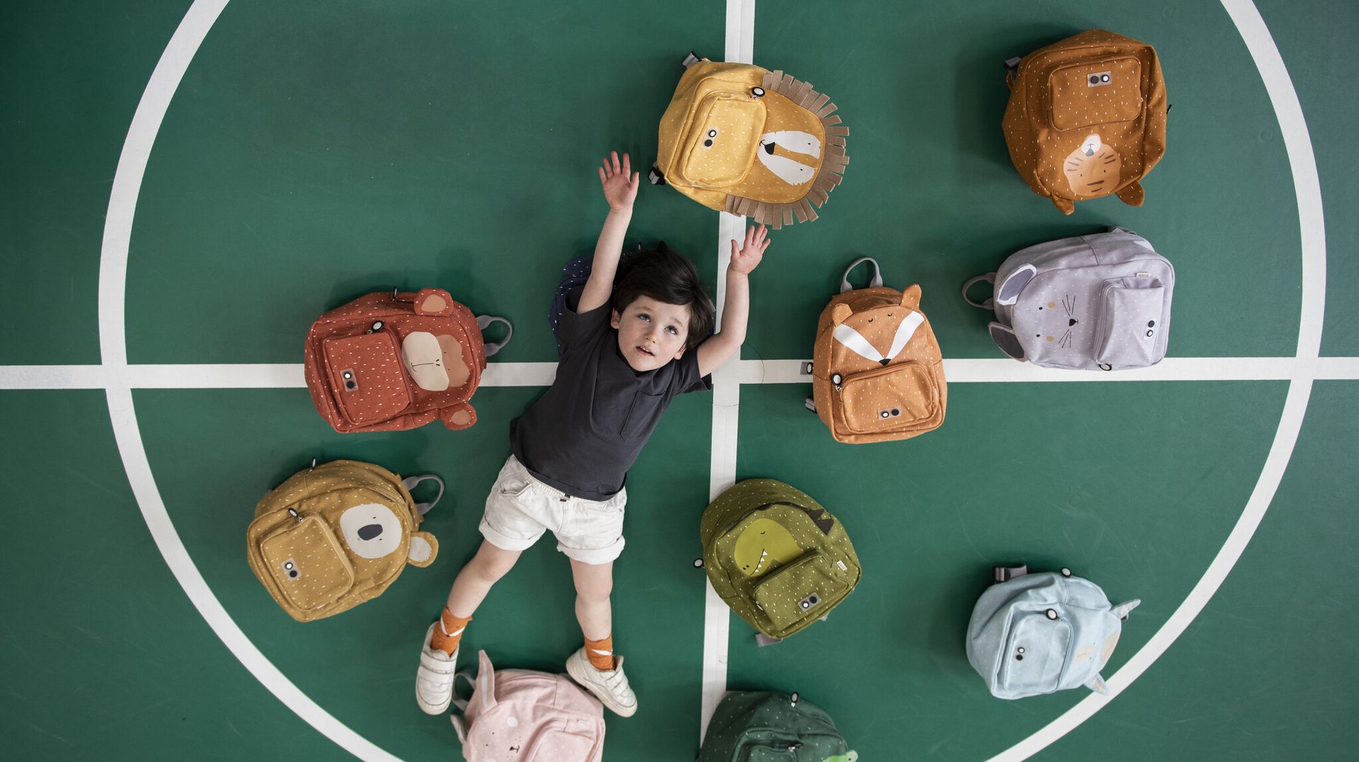 Foto in bovenaanzicht. Jongen ligt op de vloer van de turnzaal met verschillende Trixie rugzakken rond hem. Foto in bovenaanzicht. Jongen ligt op de vloer van de turnzaal met verschillende Trixie rugzakken rond hem.